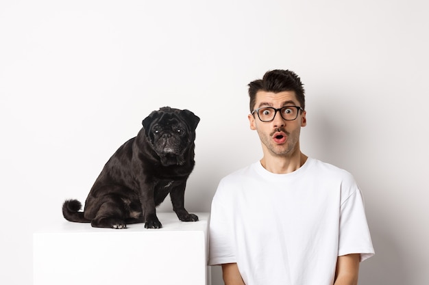 Portrait of dog owner and small cute pug staring at camera surprised and amazed, standing over white background