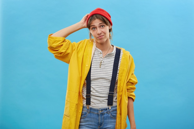 Free photo portrait of doubtful female dressed casually scratching her head with hand not knowing what to do isolated over blue wall. pretty female in yellow loose anorak being uncertain having doubts