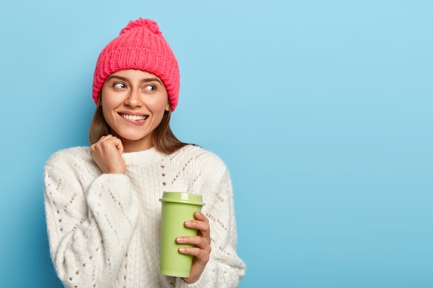 Free Photo portrait of dreamy beautiful woman gazes aside, bites lips, holds coffee cup, sees something alluring, wears pink hat and white jumper