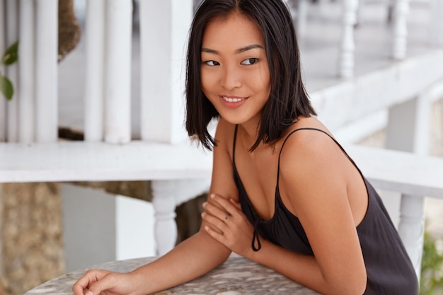 Portrait of dreamy brunette Asian female with happy expression, thinks about something, dressed in black t shirt, sits against cozy cafeteria interior. People, facial expressions and lifestyle concept