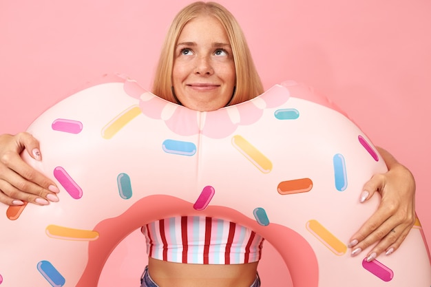 Free Photo portrait of dreamy cute blonde teenage girl in summer clothes posing isolated with pink swimming ring in shape of doughnut, looking up, dreaming about vacations by sea