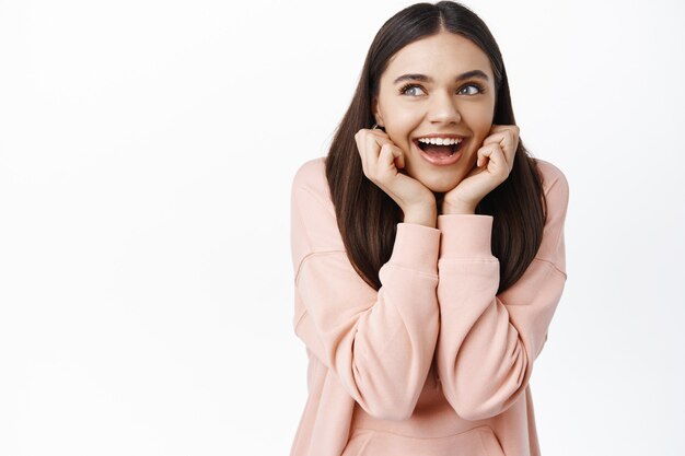 Portrait of dreamy happy woman imaging something, leaning on hands and smiling thoughtful, looking at upper left corner with interest, daydreaming, white wall