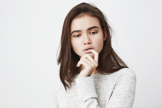 Portrait of dreamy tender woman with thoughtful expression, holding hand near mouth while biting finger and looking down