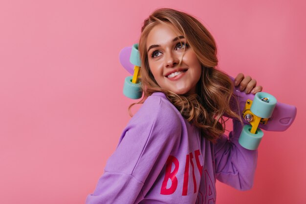 Portrait of ecstatic caucasian girl with longboard dreamy looking up. Portrait of spectacular blonde female model with pink skateboard.