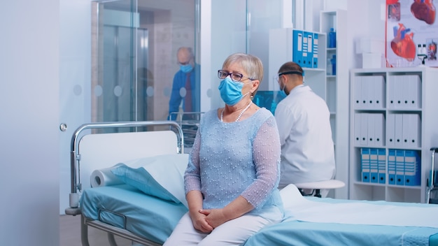 Free photo portrait of elderly lady sitting on the hospital bed during covid-19 crisis. doctor and patients wearing protective mask and equipent in modern private cinic. healthcare system after coronavirus