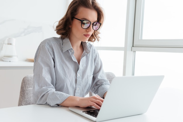 Portrait of elegant young woman texting email on laptop while sitting at table in light room