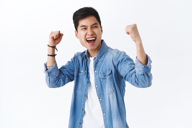 Portrait of empowered enthusiastic asian man fist pump raise hands up in triumph celebrating victory smiling saying yes winning game or competition achieve goal white background