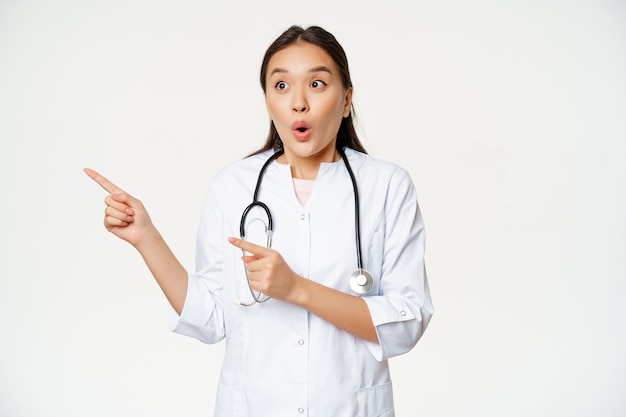 Portrait of enthusiastic female doctor, asian physician pointing and looking left with surprised, amazed face expression, standing in medical robe against white background.