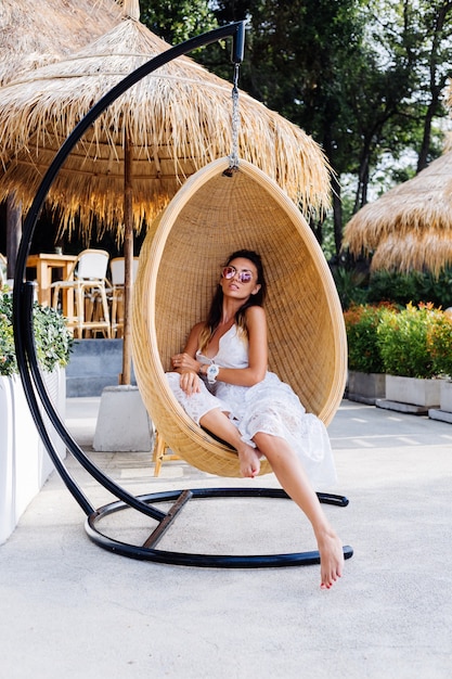 Free Photo portrait of european romantic woman in white summer dress outside hotel resort in egg straw chair.