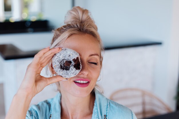 Portrait of european woman with blonde hair enjoying donuts in kitchen at home villa.