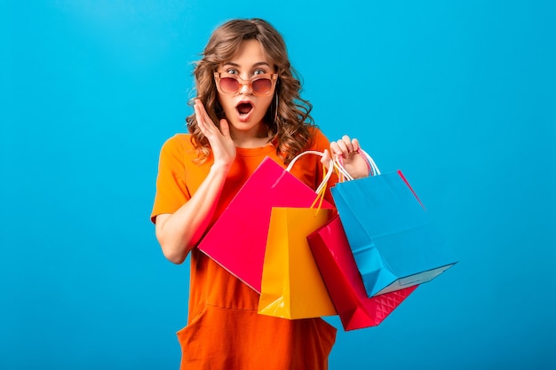 Free photo portrait of excited attractive smiling stylish woman shopaholic in orange trendy dress holding shopping bags on blue studio background isolated