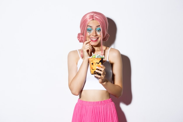 Portrait of excited beautiful girl celebrating halloween, looking at sweets with tempted expression, trick or treating in pink wig, standing.