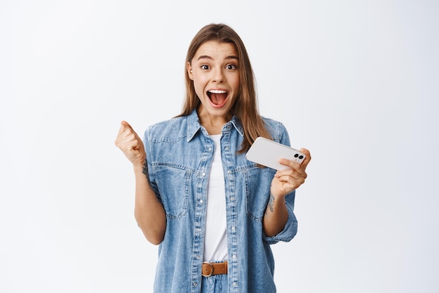 Portrait of excited blond girl winning money or prize online, holding mobile phone and screaming for joy, celebrating victory, winning in video game on smartphone, white