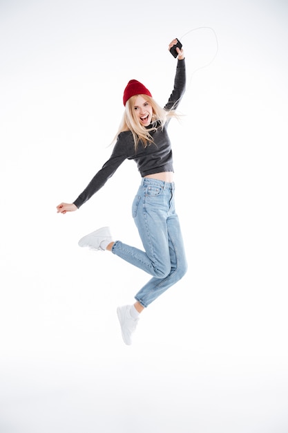 Portrait of an excited blonde young woman in hat jumping with earphones