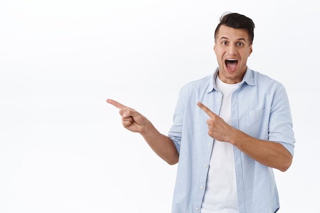 Portrait of excited, enthusiastic handsome man pointing fingers left and smiling amazed, found excellent promo, best offer in stock, special discount just click link, white wall