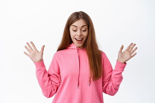 Portrait of excited gitl gasping amazed, smiling fascinated, spread hands sideways and look down with pleased face, standing over white wall