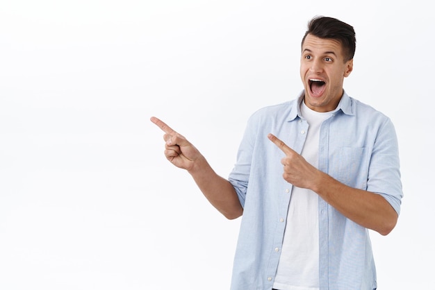 Portrait of excited happy handsome caucasian man react cheerful at incredible offer pointing fingers left and drop jaw as seeing stunning awesome new item in stock white background