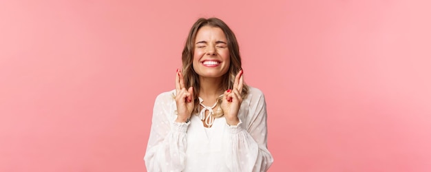 Portrait of excited hopeful blond girl making wish crossed fingers for good luck close eyes and smiling putting all effort into pray pleading for dream come true anticipating over pink background