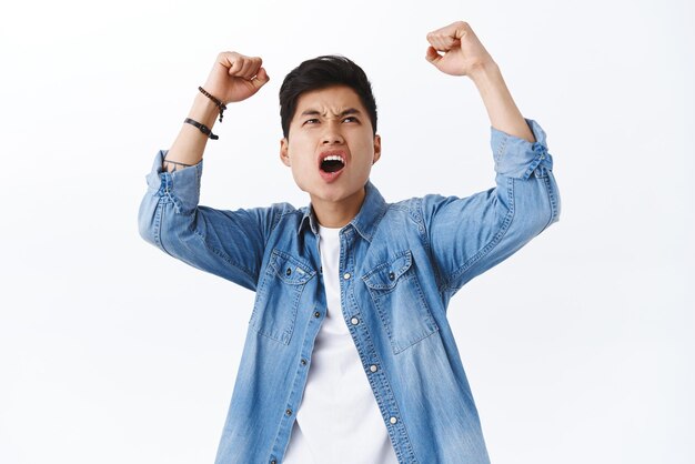 Portrait of excited intense asian man raising hands up looking at tv screen on top at pub watching sports game and triumphing fist pump over victory team scored goal white background