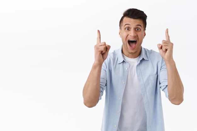 Portrait of excited joyful and impressed handsome caucasian man pointing fingers up and smiling broadly camera showing great offer super best price standing white background