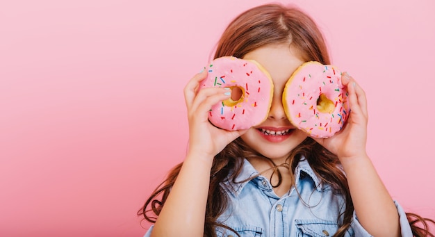 Free photo portrait excited joyful young pretty girl in blue shirt expressing positivity, having fun to camera with donuts on eyes isolated on pink background. happy childhood with tasty dessert. place fot text