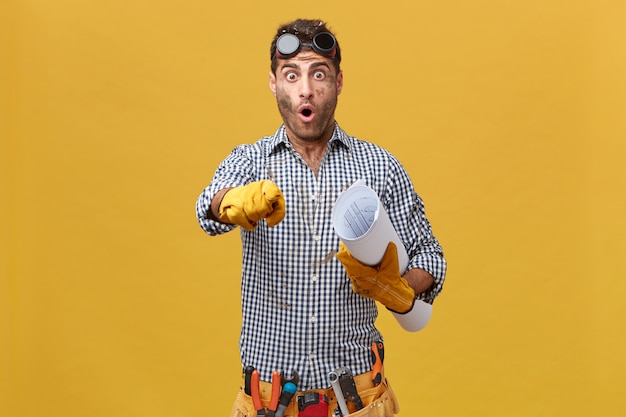 Portrait of excited male plumber wearing protective eyeglasses, checkered shirt, belt with instruments holding paper in hand pointing with forefinger. Professional workman looking puzzled