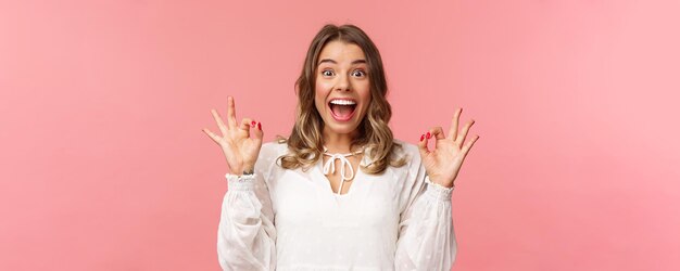 Portrait of excited and upbeat cheerful blond girl in white dress dancing and showing okay signs with happy beaming smile excited over music festival rate awesome movie recommend product