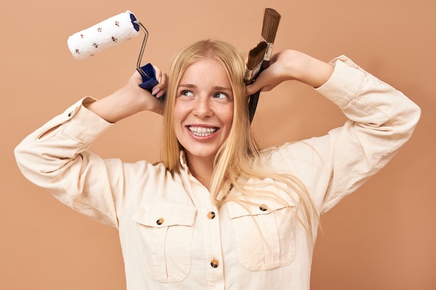 Portrait of excited young Caucasian woman holding paintbrush and roller while painting walls in her bedroom