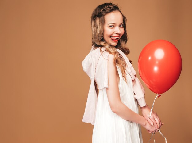 Portrait of excited young girl posing in trendy summer white dress. Smiling woman with red balloon posing. Model ready for party, showing her tongue 