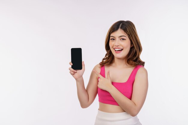 Portrait of excited young woman holding mobile phone mock up and pointing finger at smartphone isolated over white background