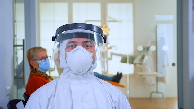 Portrait of exhausted doctor in dental office looking on camera wearing ppe suit and face shield sitting on chair in waiting room clinic. Concept of new normal dentist visit in coronavirus outbreak.