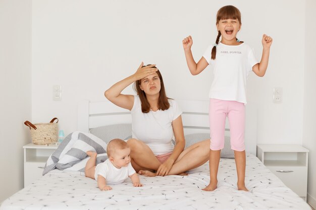 Portrait of exhausted female wearing white t shirt sitting on bed with her two loud daughters, keeping hand on forehead, being tired to spend lots time with children.