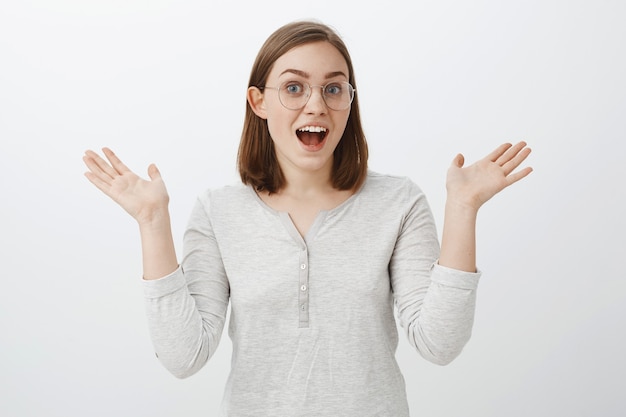 Free photo portrait of expressive joyful smart european female in trendy glasses and white blouse talking passionately about interesting hobby gesturing with raised palms being emotional over grey wall