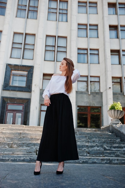 Portrait of a fabulous young successful woman in white blouse and broad black pants posing on the stairs with a huge white building on the background