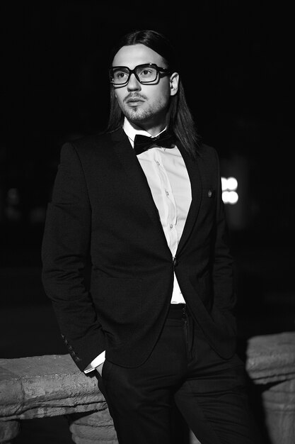 Portrait of fashion elegant long-haired young man. Attractive and handsome male model in black suit with mustache in the street at night