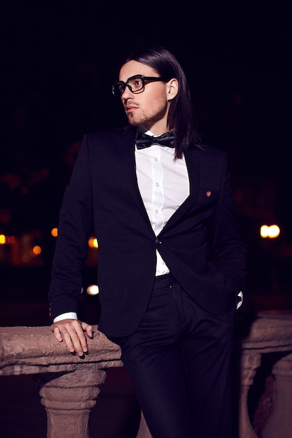 Portrait of fashion elegant long-haired young man. Attractive and handsome male model in black suit with mustache in the street at night