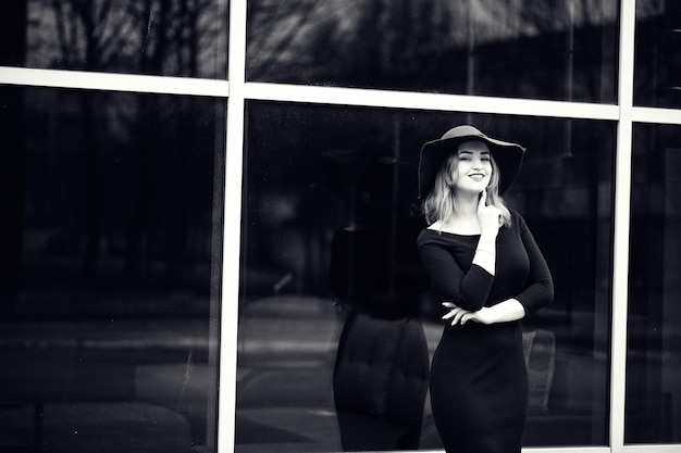 Portrait of fashion red haired girl on red hat and black dress with bright make up posed against large window Black and white photo