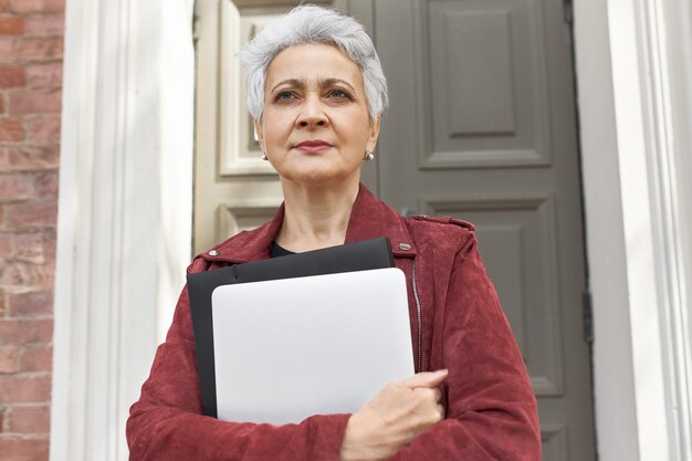 Portrait of fashionable 50 year old Caucasian woman holding portable computer posing outside her house