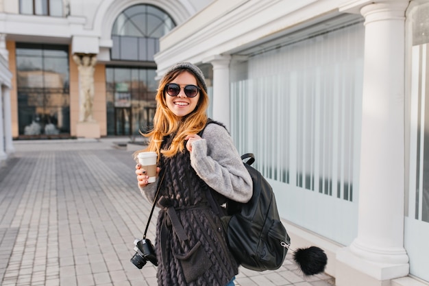 Free Photo portrait fashionable attractive woman walking with coffee to go in city centre. young smiling stylish woman in modern sunglasses, winter sweater, knitted hat travelling with bag, camera.