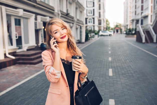 Portrait fashionable blonde woman with long hair walking in coral jacket on street. She is speaking on phone, holds a cup