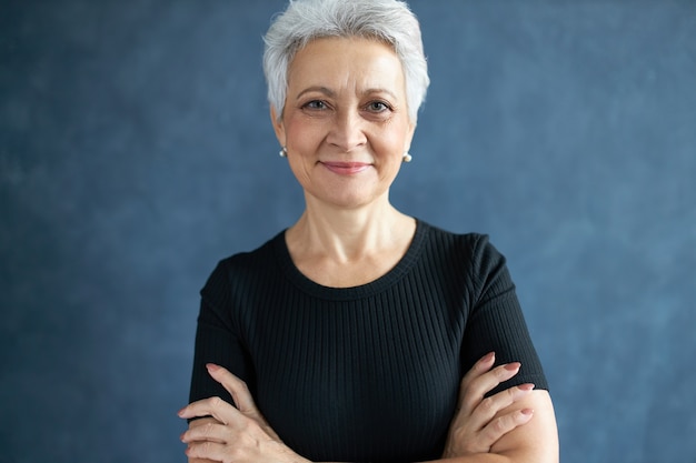 Free photo portrait of fashionable gray haired female on retirement posing isolated in black t-shirt keeping arms crossed, being in good mood, looking at camera with confident smile, expressing positive emotions