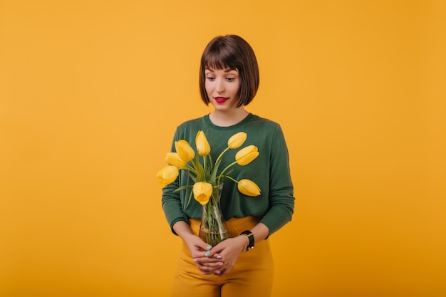 Free photo portrait of fashionable lady with trendy haircut holding vase of tulips. indoor photo of interested brunette girl isolated with flower bouquet.