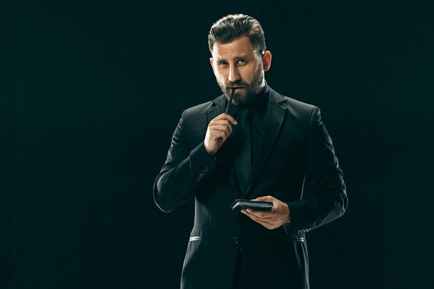 Portrait of a fashionable young man with stylish haircut wearing trendy suit posing over black wall