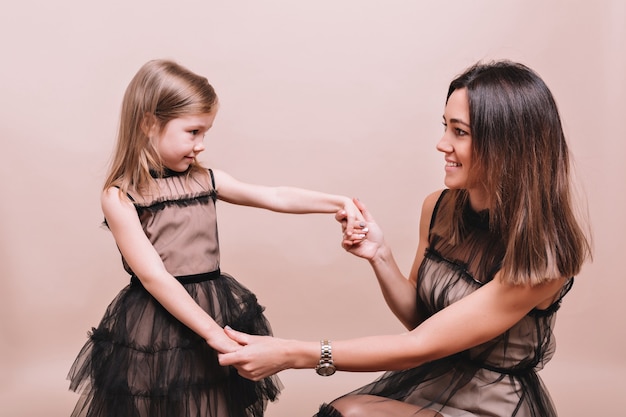 Portrait of fashionable young woman with little cute girl wearing similar black dresses posing on beige wall with truly emotions. Stylish family look of mother and daughter