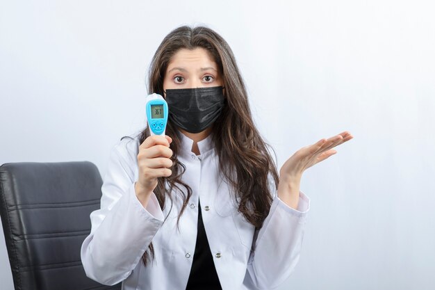 Portrait of female doctor in medical mask and white coat holding thermometer. 