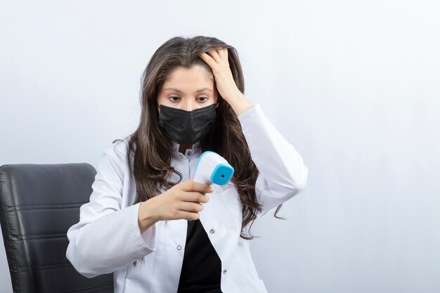 Portrait of female doctor in medical mask and white coat looking at thermometer.