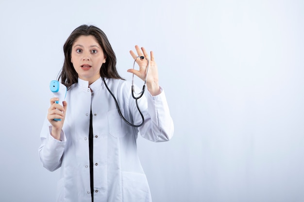 Portrait of female doctor showing thermometer and stethoscope on grey. 