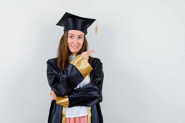 Portrait of female graduate pointing right in academic dress and looking hopeful front view