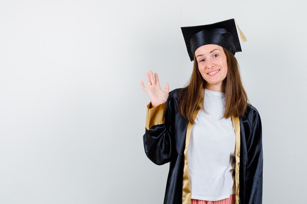 Portrait of female graduate waving hand for greeting in academic dress and looking merry front view