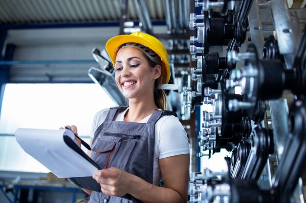 Portrait of female industrial employee in working uniform and hardhat writing production results in factory